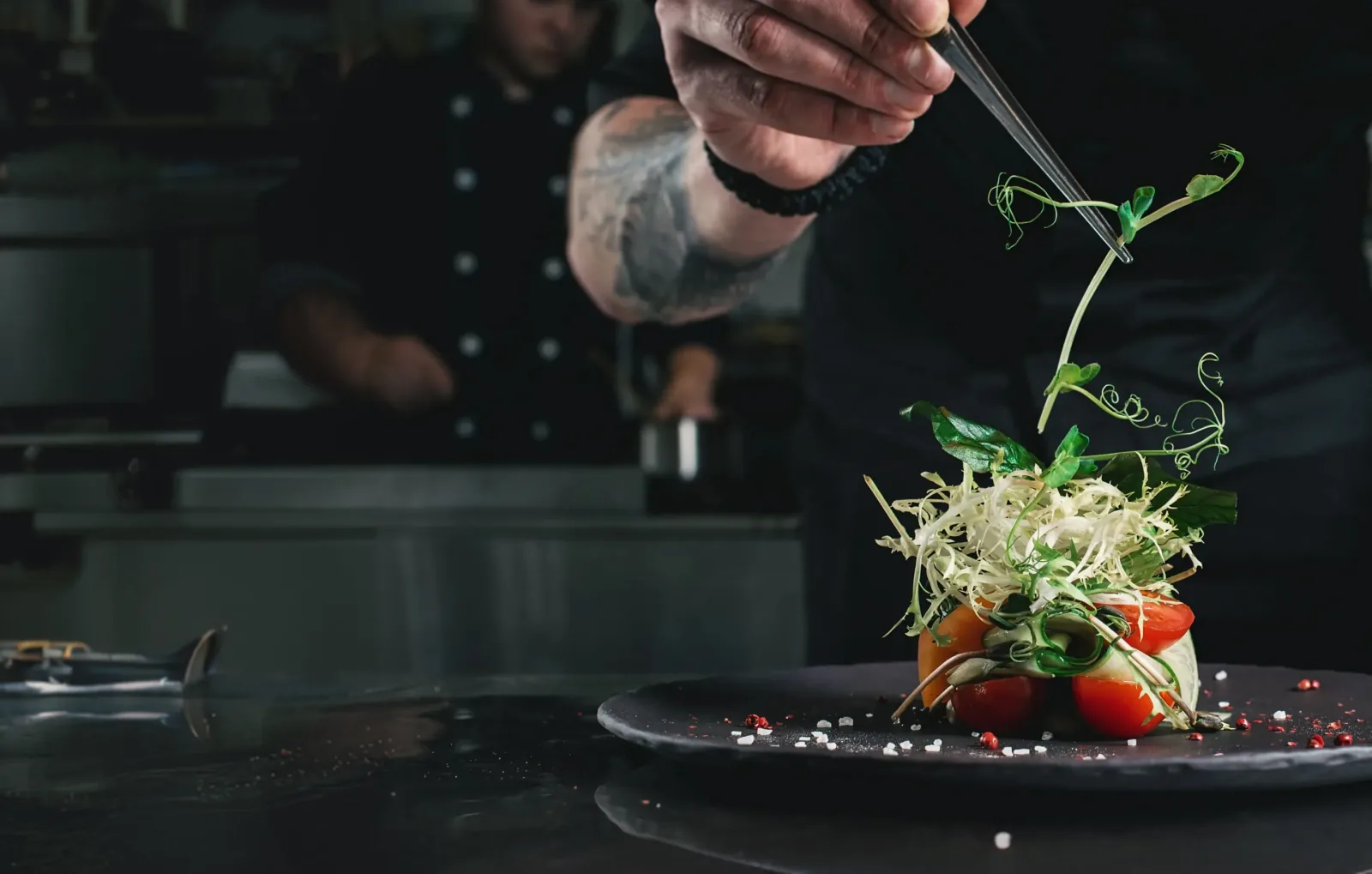 A chef garnishes a plated salad of tomato and frisée with microgreens using plating tweezers