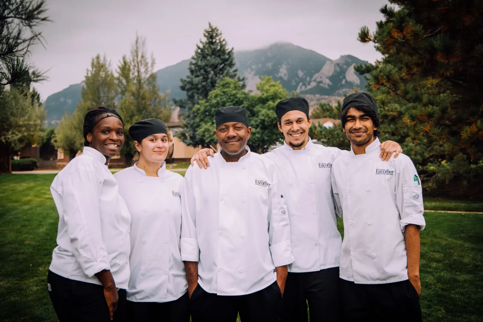 Five Escoffier culinary students in chef whites stand arm in arm on the Boulder campus lawn, mountain foothills behind them