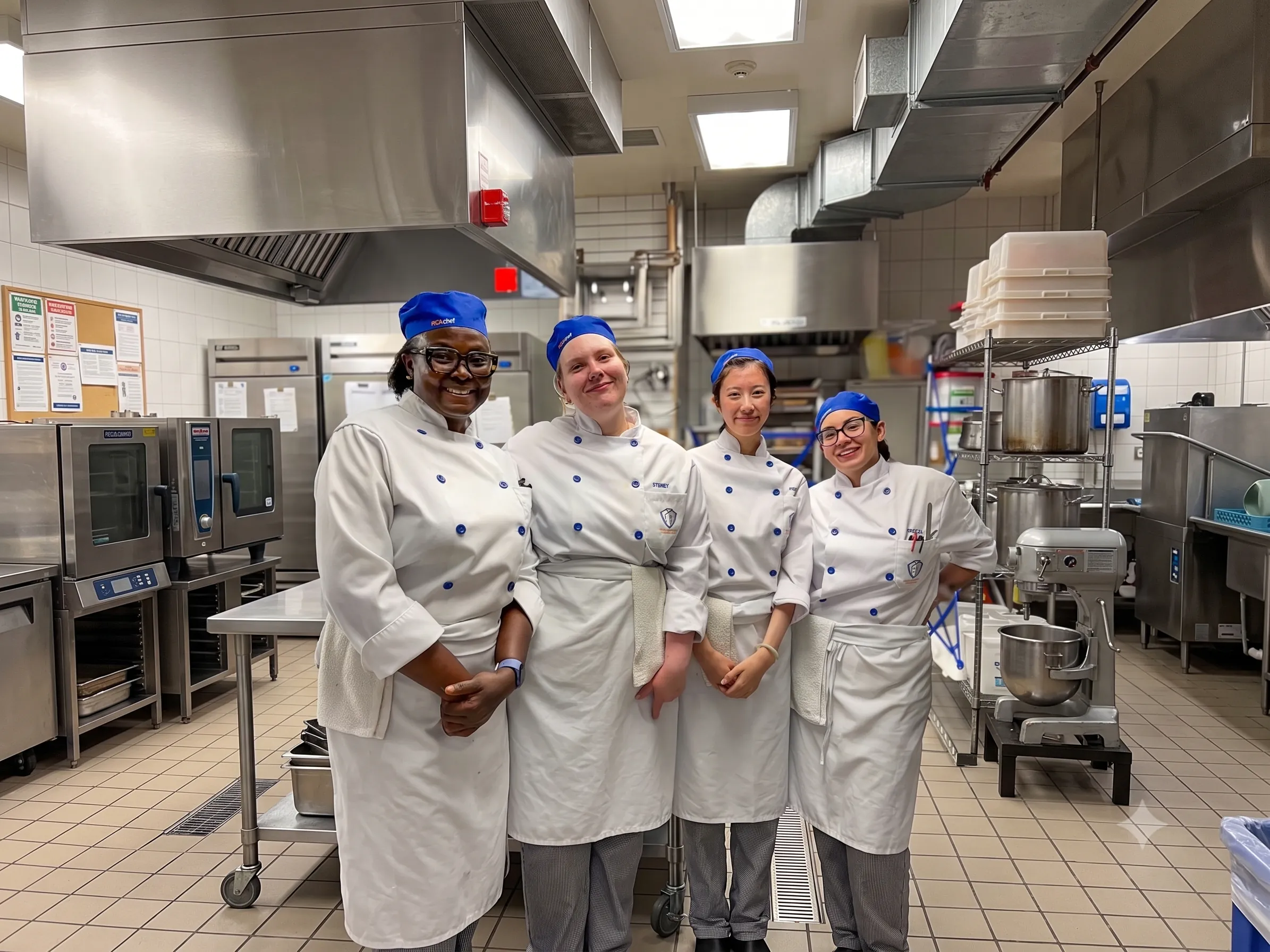 Four PICAchef culinary students in blue caps and chef whites smile together in a teaching kitchen