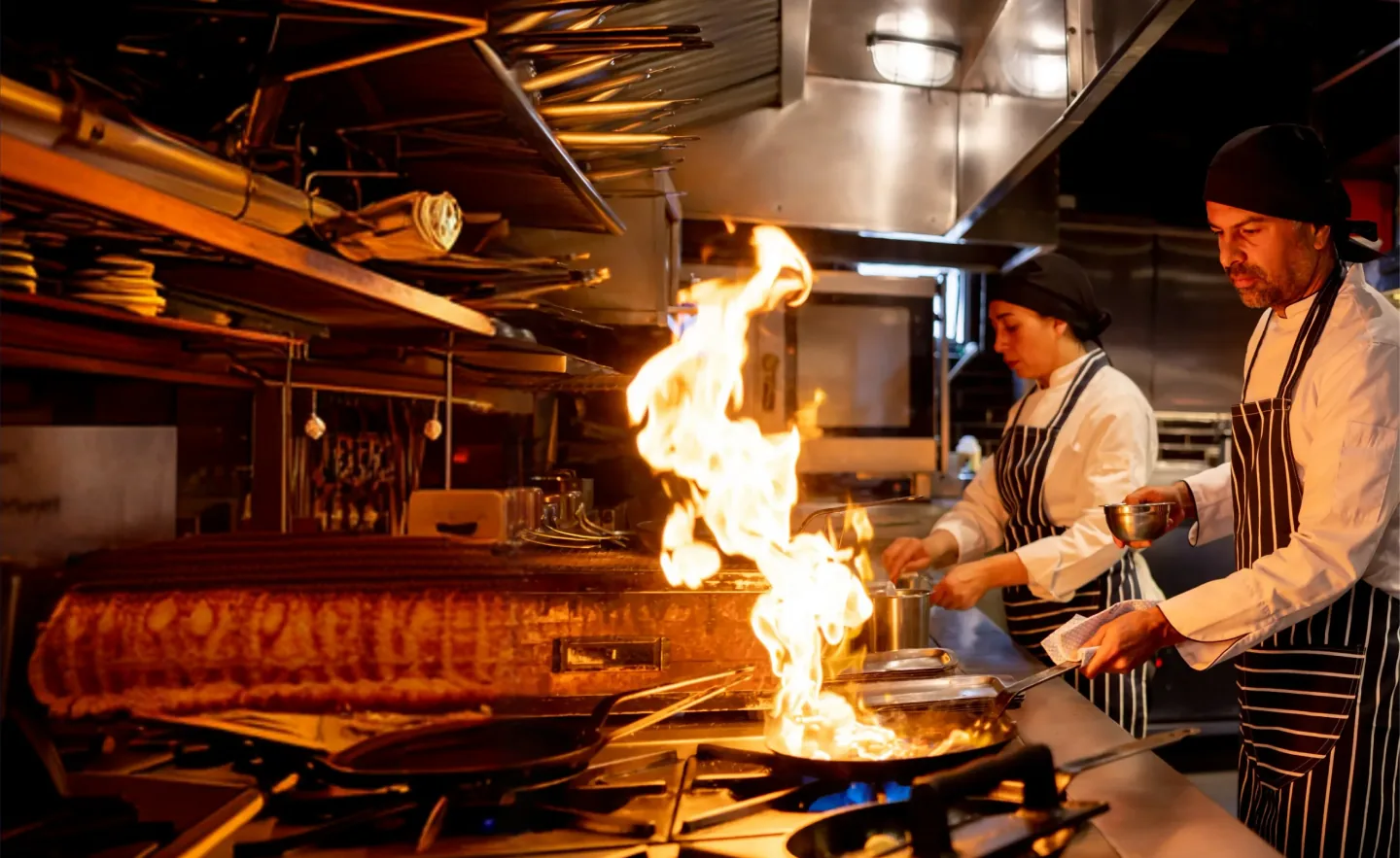 Two chefs work the line in a restaurant kitchen as flames rise from a saut&eacute; pan